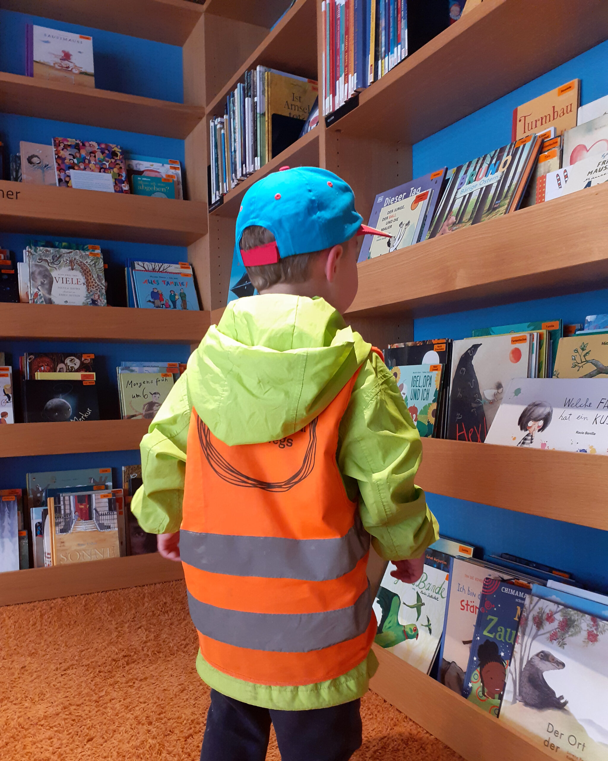 Blick von hinten auf ein Kind mit einer grünen Jacke und einer orangenen Warnweste. Das Kind betrachtet die Bücher in einem Bücherregal der Bücherei. (c) KibeV