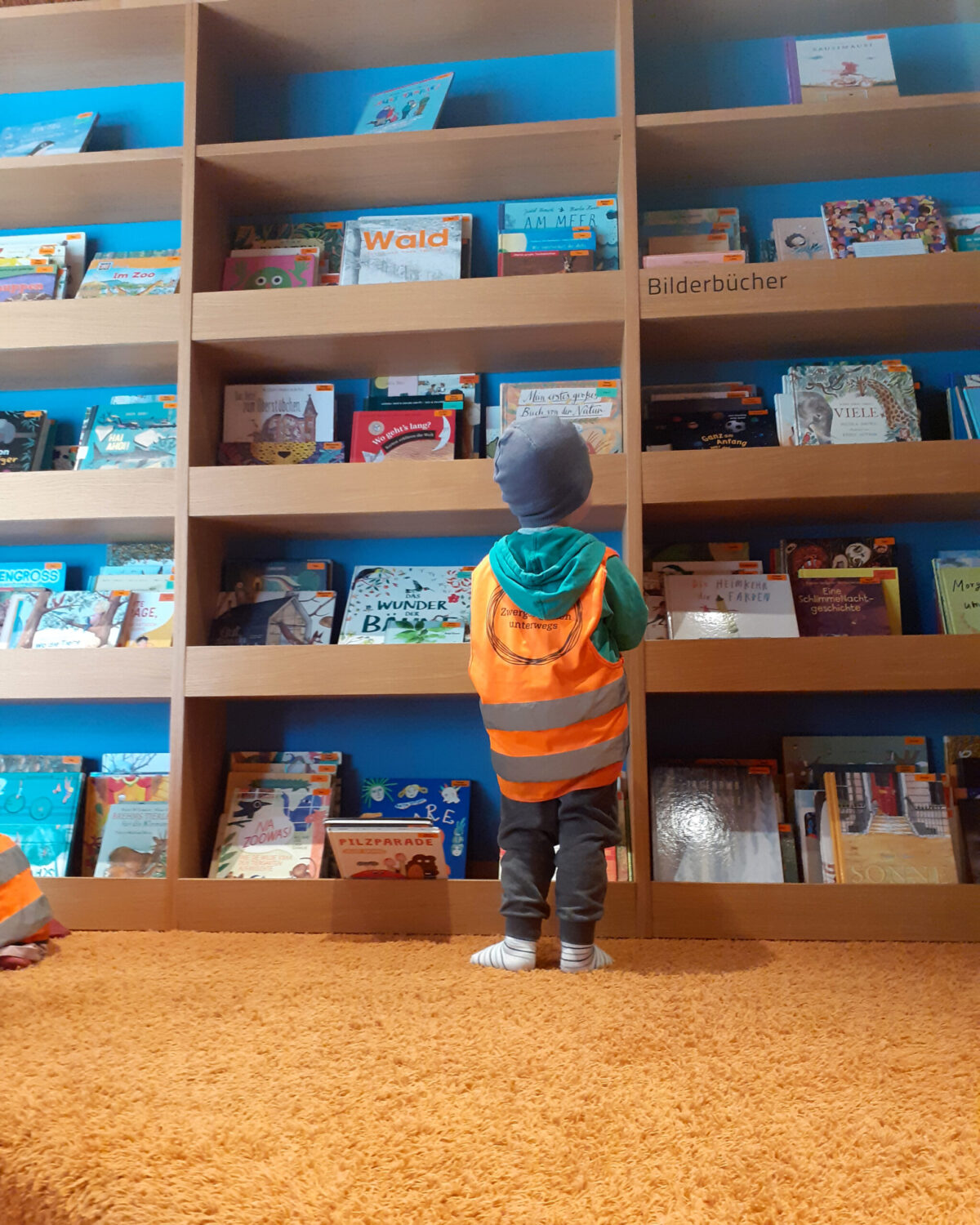 Blick von hinten auf ein Kind, das vor dem riesigen Bücherregal steht und sich ein Buch aussucht. Das Kind trägt eine blaue Jacke und eine orangene Warnweste. (c) KibeV