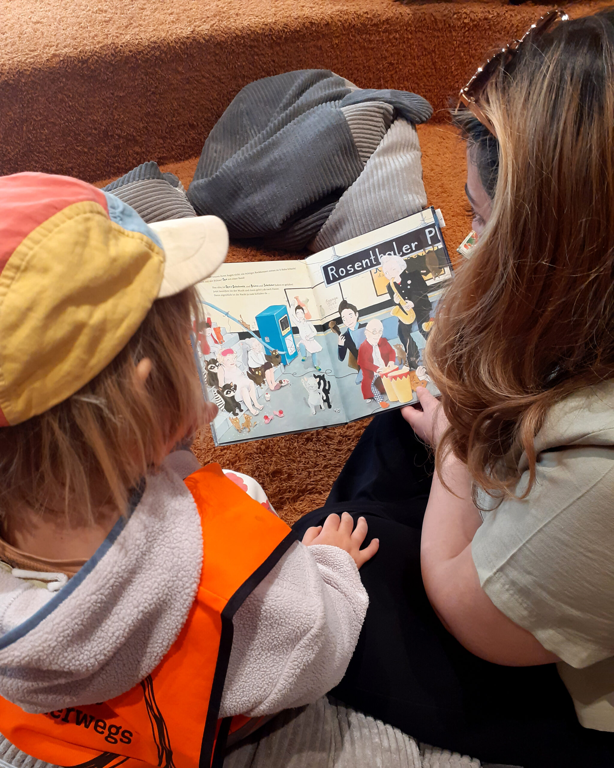 Ein Kind und eine Betreuerin sitzen auf dem Boden und betrachten eine bunte Seite eines Kinderbuchs. (c) KibeV