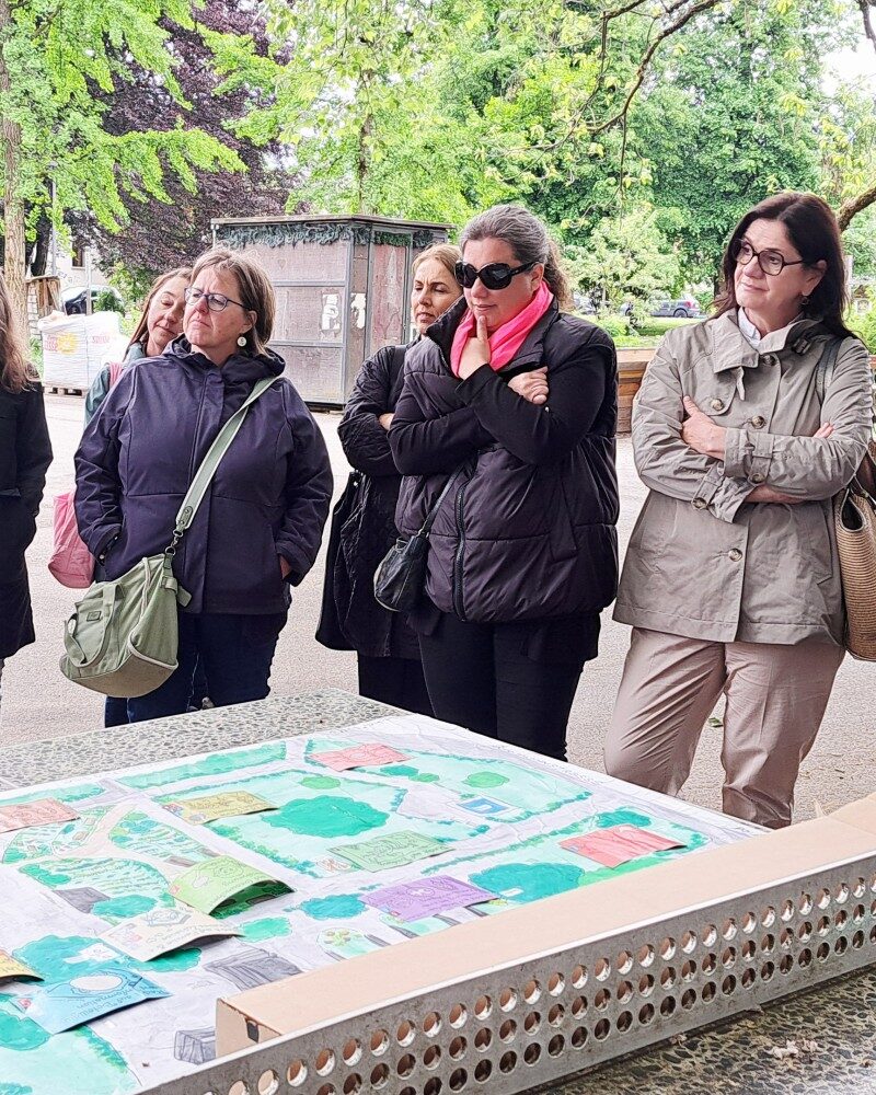 Unsere Regionalleitungen stehen um Nico Kantner (Jugendbotschafter:innen Vorarlberg) und erfahren alles über den Kinderrechteplatz Vorarlberg. Auf dem Tischtennnistisch vor ihnen liegt ein Plan des Platzes - Teil 2. (c) KibeV
