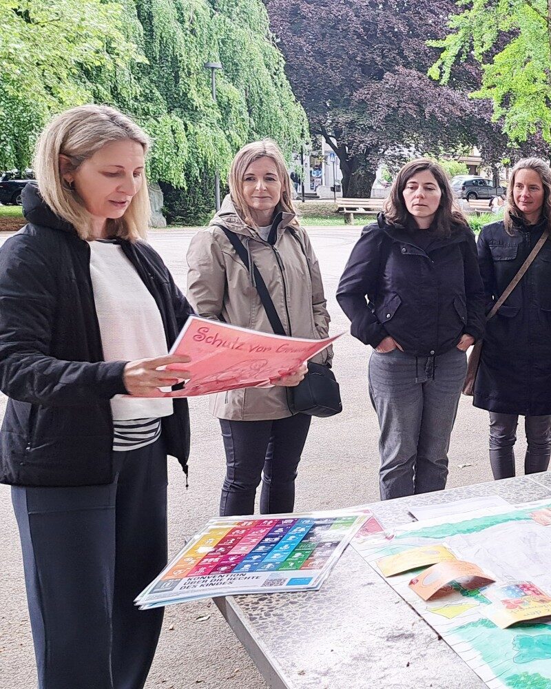 Unsere Regionalleitungen stehen um Nico Kantner (Jugendbotschafter:innen Vorarlberg) und erfahren alles über den Kinderrechteplatz Vorarlberg. Auf dem Tischtennnistisch vor ihnen liegt ein Plan des Platzes - Teil 1. (c) KibeV