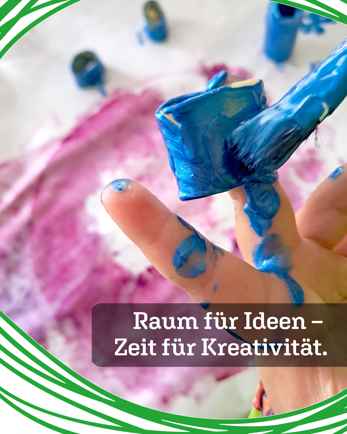 Close-up of a finger smeared with bright blue paint, with a pink-painted background and a green decorative border, caption reads 'Raum für Ideen – Zeit für Kreativität'