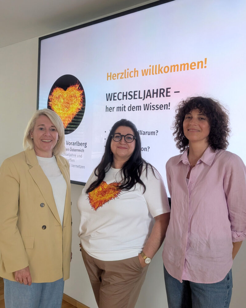 Drei Frauen stehen lächelnd vor einem hellen Banner mit Willkommensgruß und Informationen zum Thema Wechseljahre. Bannertext: 'Herzlich willkommen! WECHSELJAHRE – hier mit dem Wissen!'