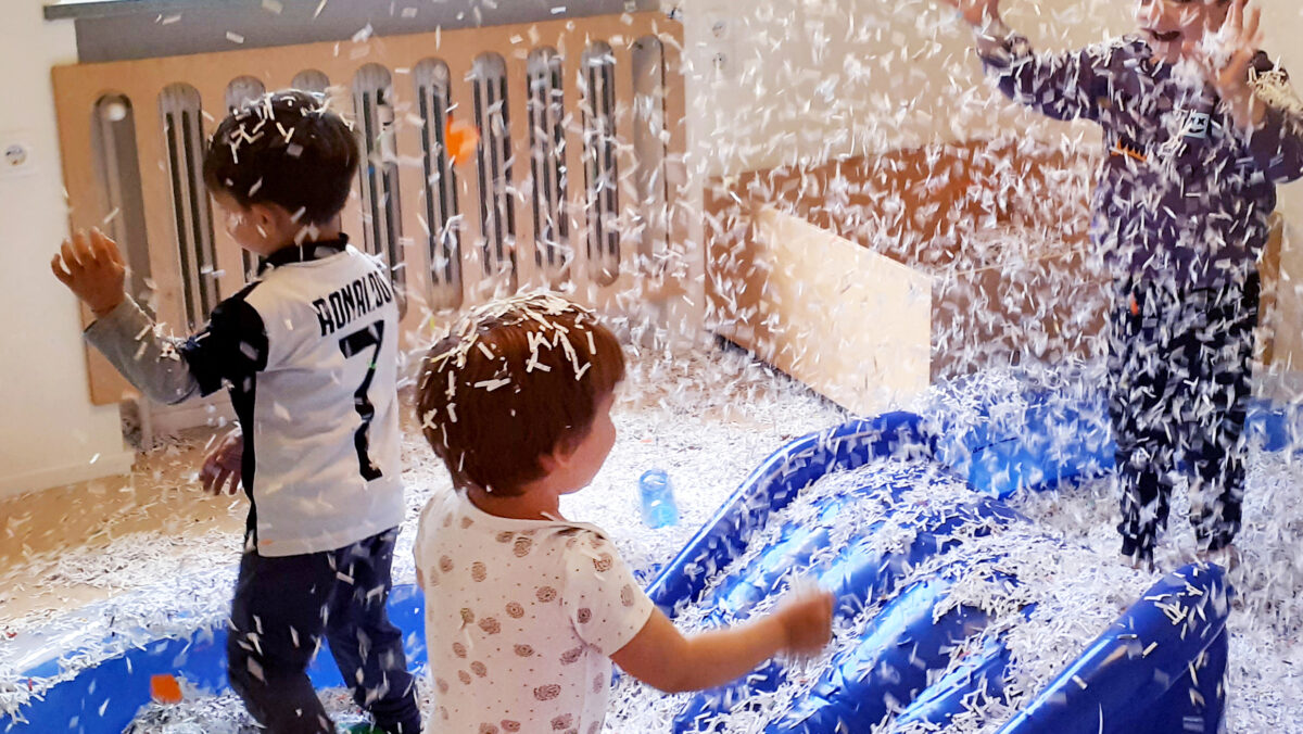 Aktion "PARKplatz": 3 Kinder im Zwergengarten Neuner spielen in einem blauen Schwimmbecken mit Rutsche mit Papierschnipseln. (c) KibeV