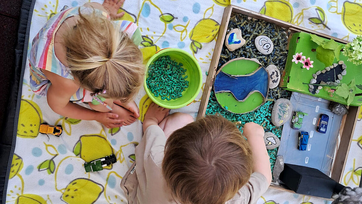 2 Kinder sitzen auf einer weißen Decke mit aufgedruckten gelben Zitronen. Sie streuen grüne Nudeln in ihre Sensory-Box mit dem Thema "ökologischer Fußabdruck". (c) KibeV