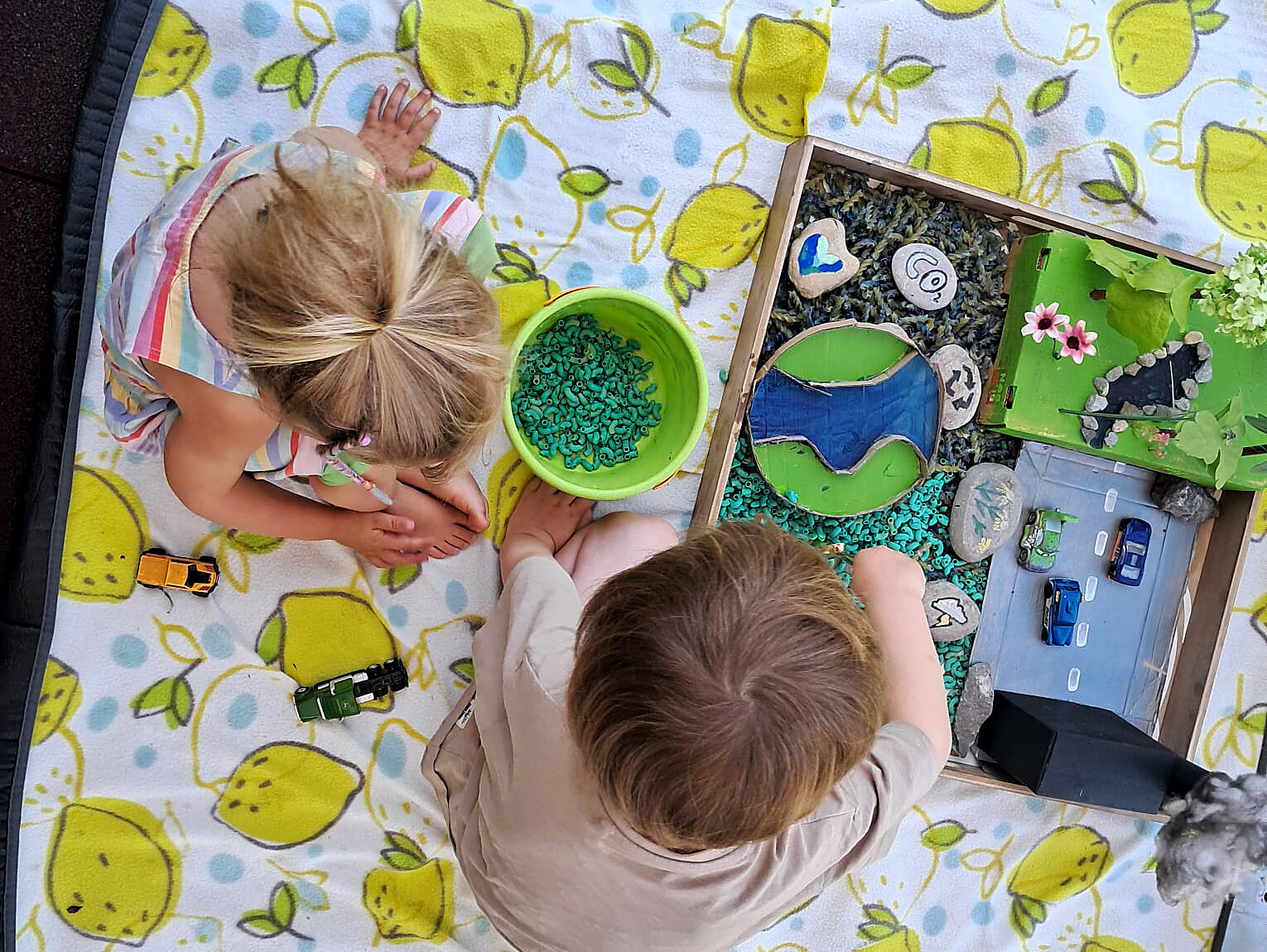 2 Kinder sitzen auf einer weißen Decke mit aufgedruckten gelben Zitronen. Sie streuen grüne Nudeln in ihre Sensory-Box mit dem Thema "ökologischer Fußabdruck". (c) KibeV