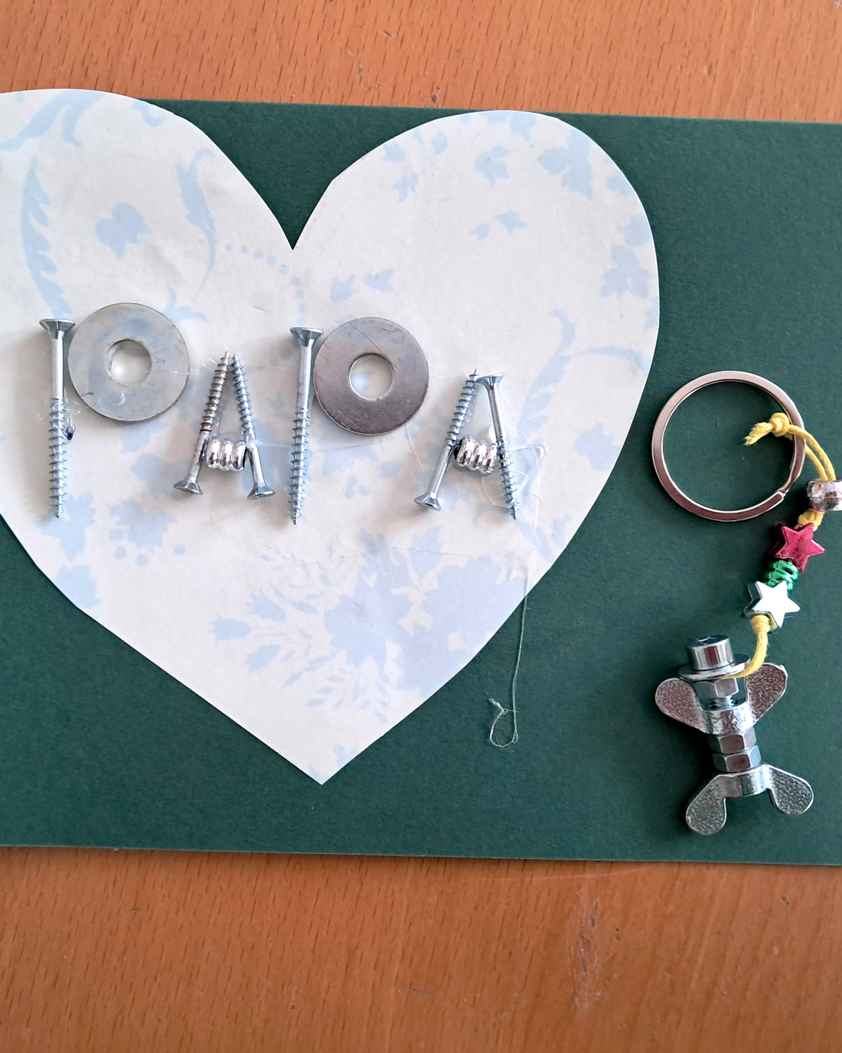 Vatertagskarte aus grünem Papier mit weiß-blau-marmorierten Herz darauf. Im Herz steht "Papa" geformt aus Schrauben, Muttern und Unterlegscheiben Daneben ist ein Schlüsselanhänger angebracht. Dieser besteht aus einer Schraube und jeweils 3 Muttern sowie 2 Flügelschrauben, die darauf geschraubt wurden. (c) KibeV