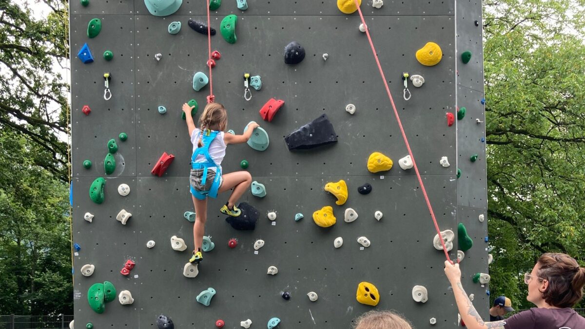 Ein Kind mit einer blauen Hose und einem Pinken Shirt kletter eine Kletterwand mit bunten Haltegriffen hoch. Es ist durch ein Seil gesichert. (c) KibeV