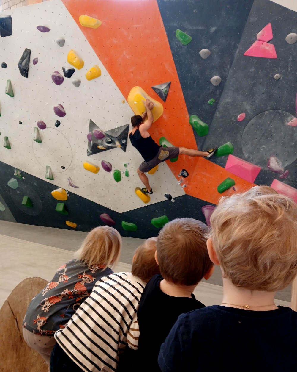 Climber in a black tank top scales a colorful indoor bouldering wall while several children watch from below.