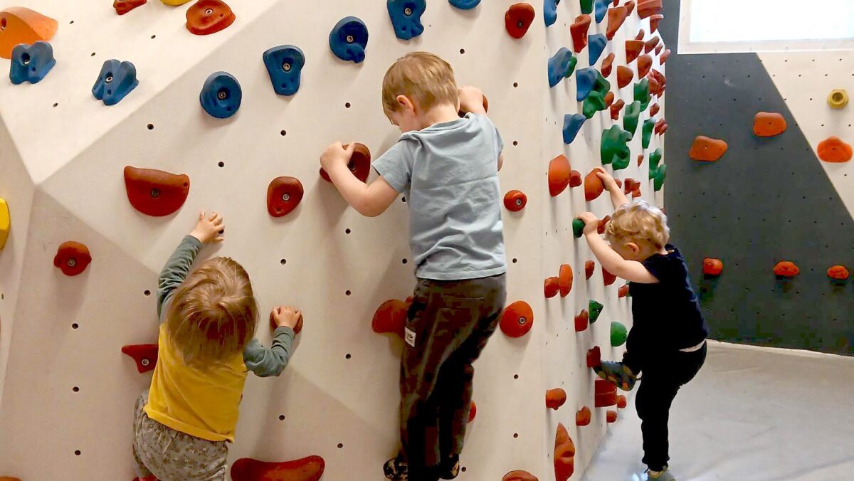 Drei Kleinkinder versuchen, eine niedrige Boulderwand hochzuklettern. (c) KibeV