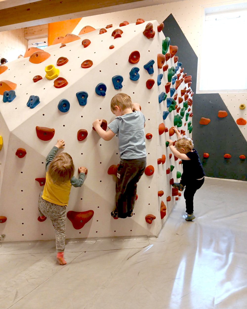 Three children climb a large white indoor climbing wall with red, blue, and green holds in a gym.