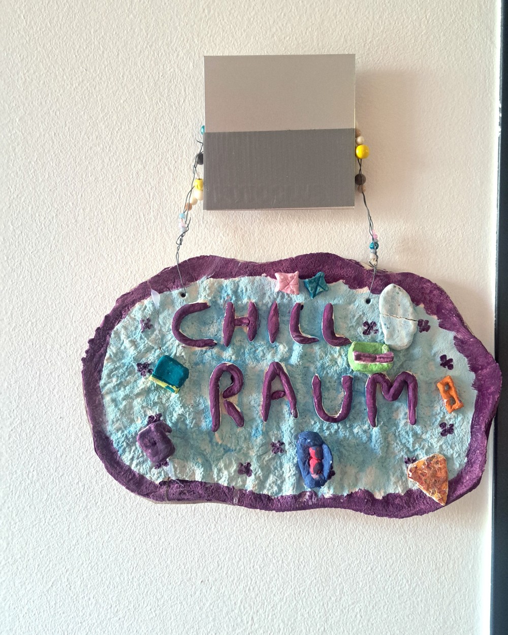 Handmade sign reading 'CHILL RAUM' in purple letters on light blue with a purple border, hanging by a bead string above.