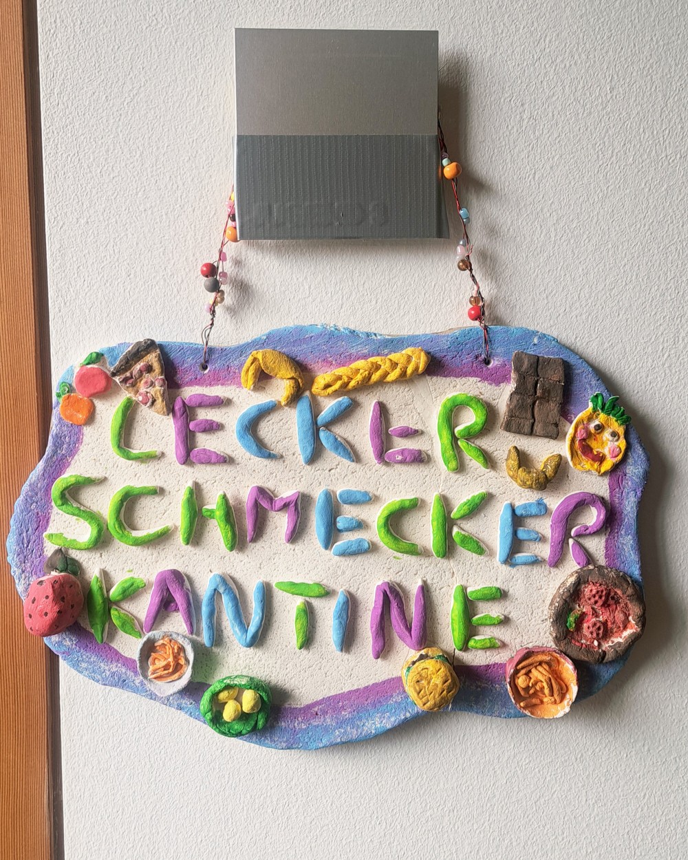 Colorful clay sign reading 'Lecker schmecker Kantine' with toy food decorations hanging on a wall.