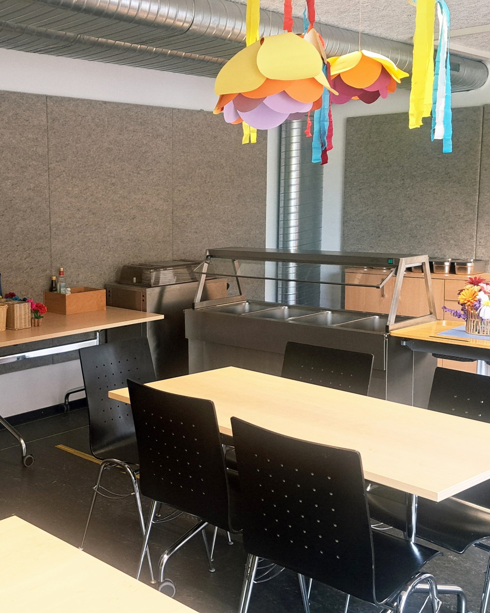 Cafeteria-style dining area with light wood tables, black chairs, and colorful paper lanterns hanging from the ceiling to create a festive look.