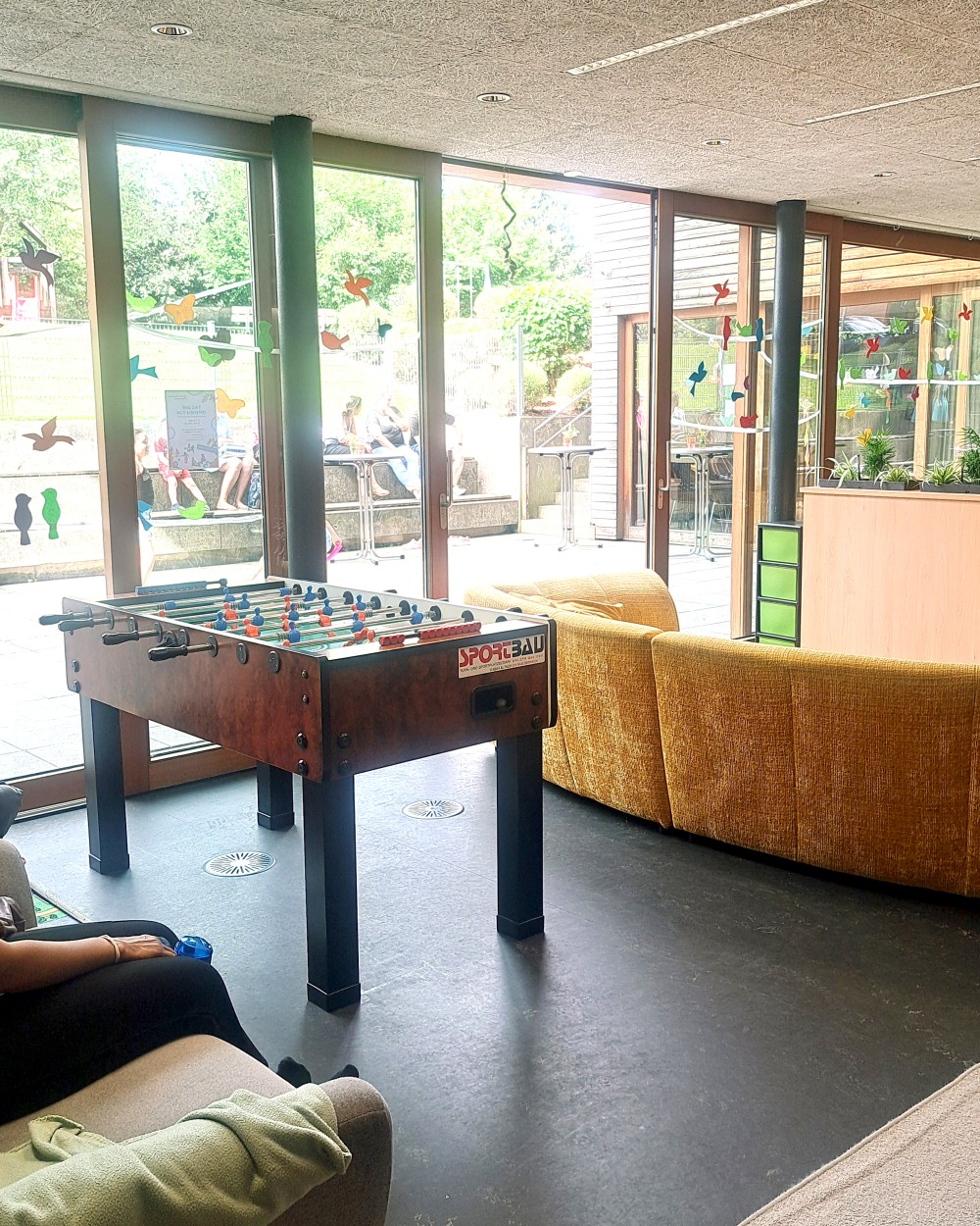 Foosball table in a bright lounge with a yellow sofa and large glass doors opening to an outdoor seating area.