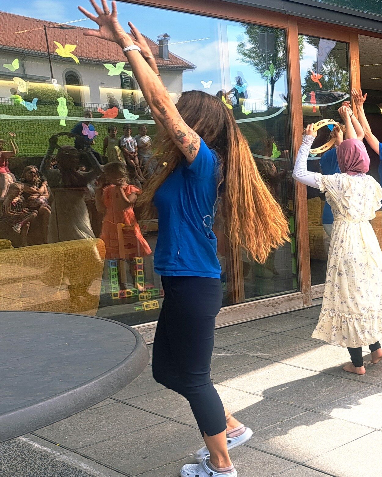 Group of children and adults outside a glass-front building, raising hands as part of an outdoor activity or dance.