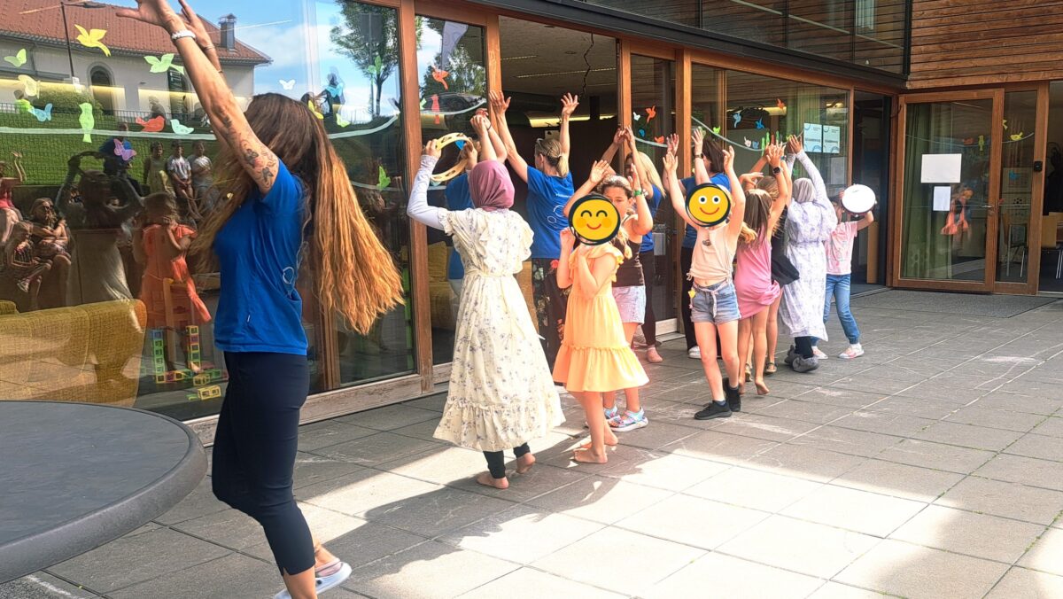 Children and adults with hands raised outside a glass-front building during a sunny group activity, reflections visible in the windows.
