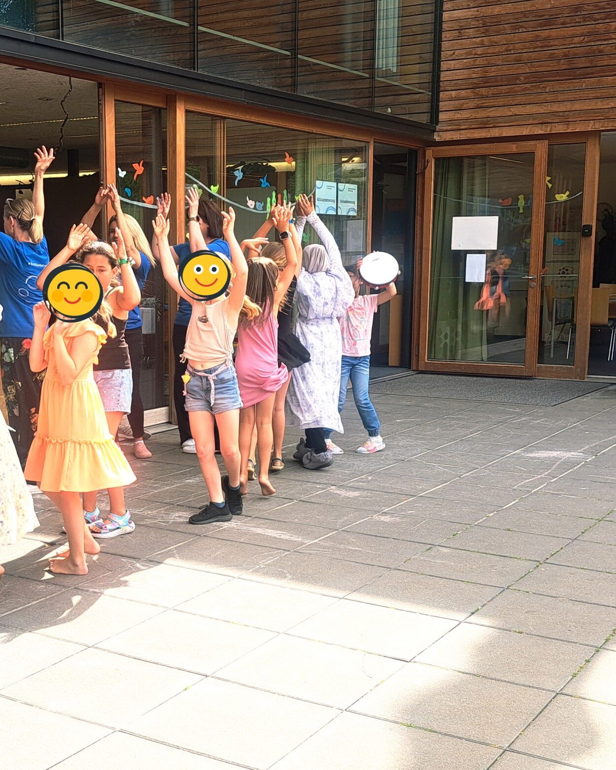 Group of children outside a glass-front building, raising hands and dancing in a playful outdoor activity, supervised by adults nearby.