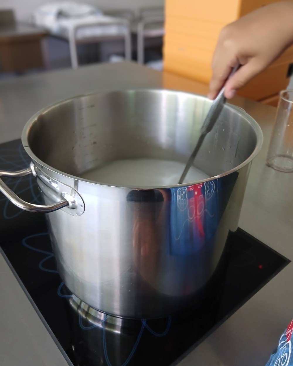 Hand stirring a white liquid in a large stainless steel pot on an induction cooktop.