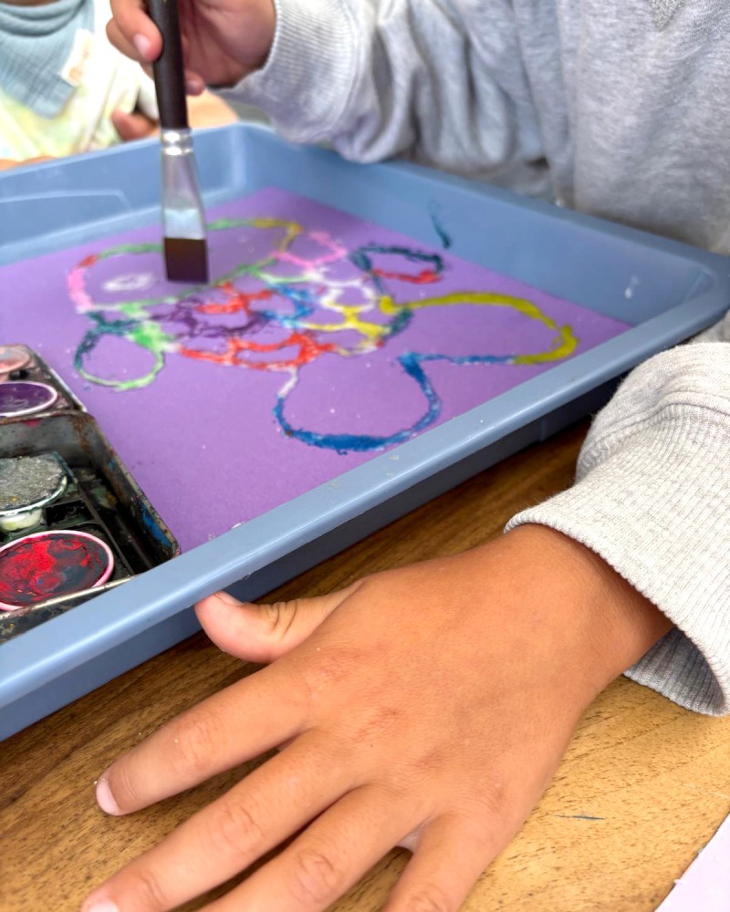 Blick auf ein Tablett mit Umrissen eines Fisches. Kinderhände halten einen Pinsel und bemalen dies Umrisse mit bunten Farben. (c) KibeV