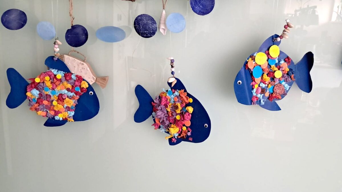 Drei Regenbogenfische aus Papier sind an einem Holzast an einer Wand befestigt. (c) KibeV