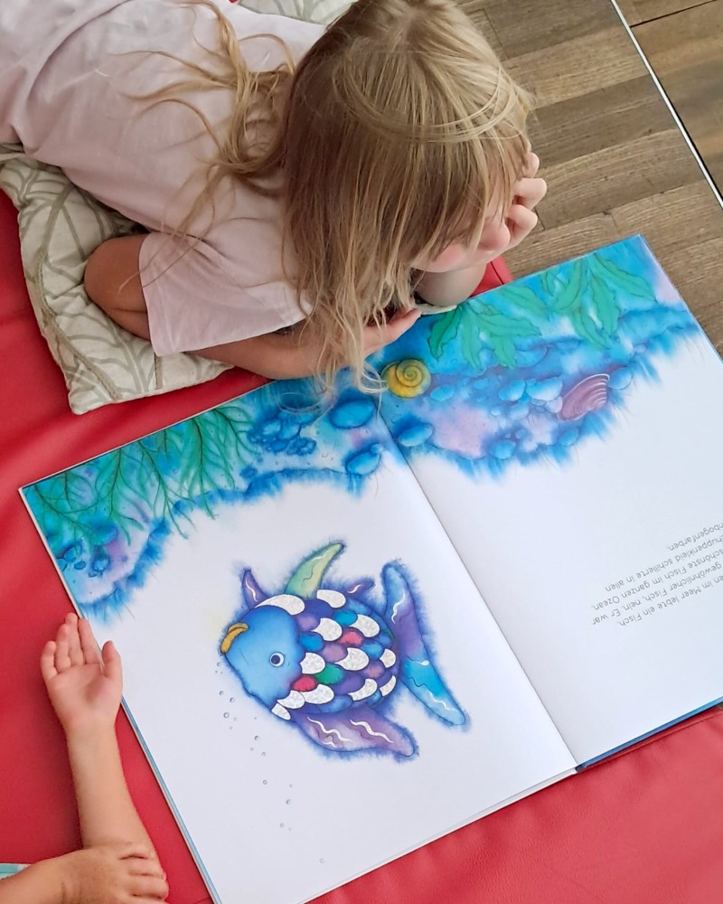 Blick von oben auf ein Kind, das auf einer Matte liegt und das Buch "Regenbogenfisch" liest. (c) KibeV