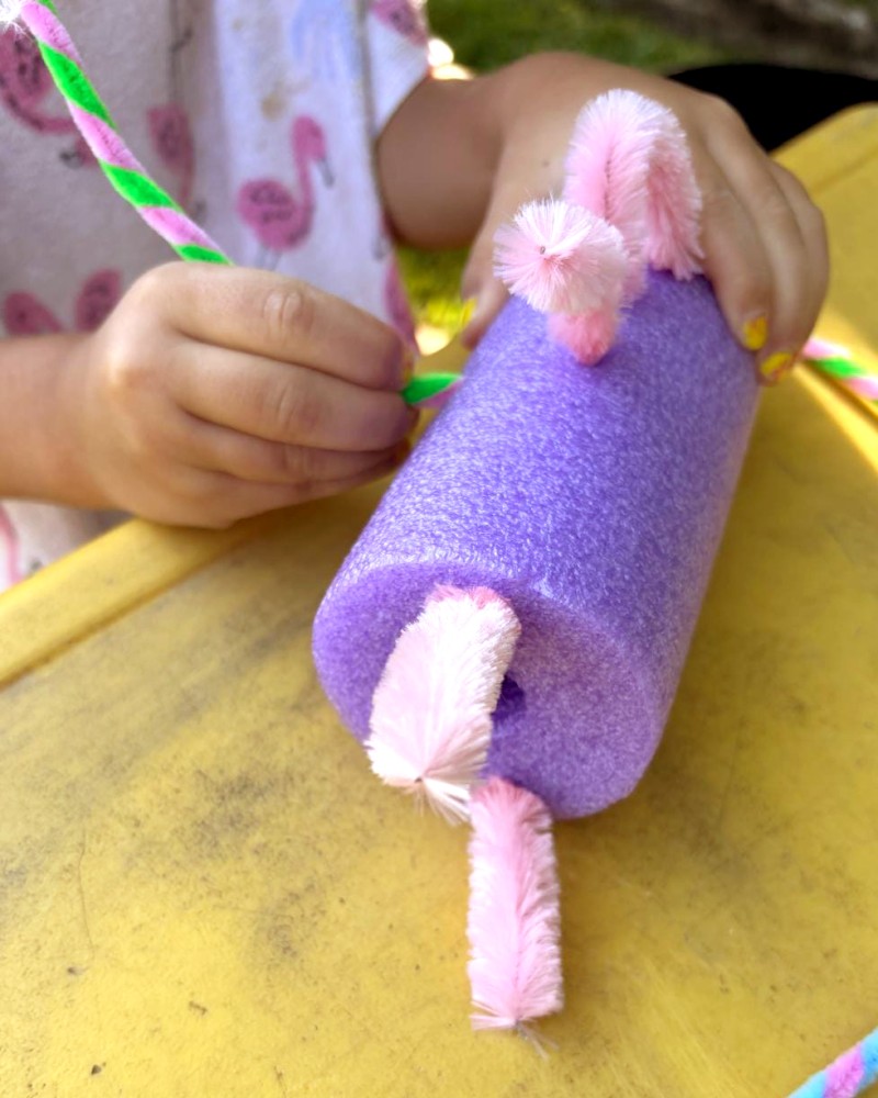 Closeup auf zwei Kinderhände, die einen Regenbogenfisch aus einer lila Poolnudel mit rosa Pfeifenputzern basteln. (c) KibeV