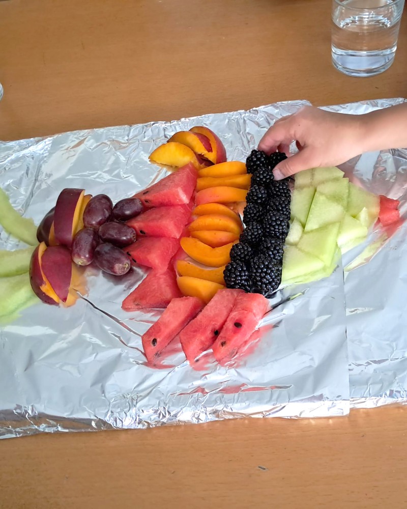 Eine Kinderhand nimmt sich eine Brombeere von einer Obstplatte in Form eines Regenbogenfisches. (c) KibeV