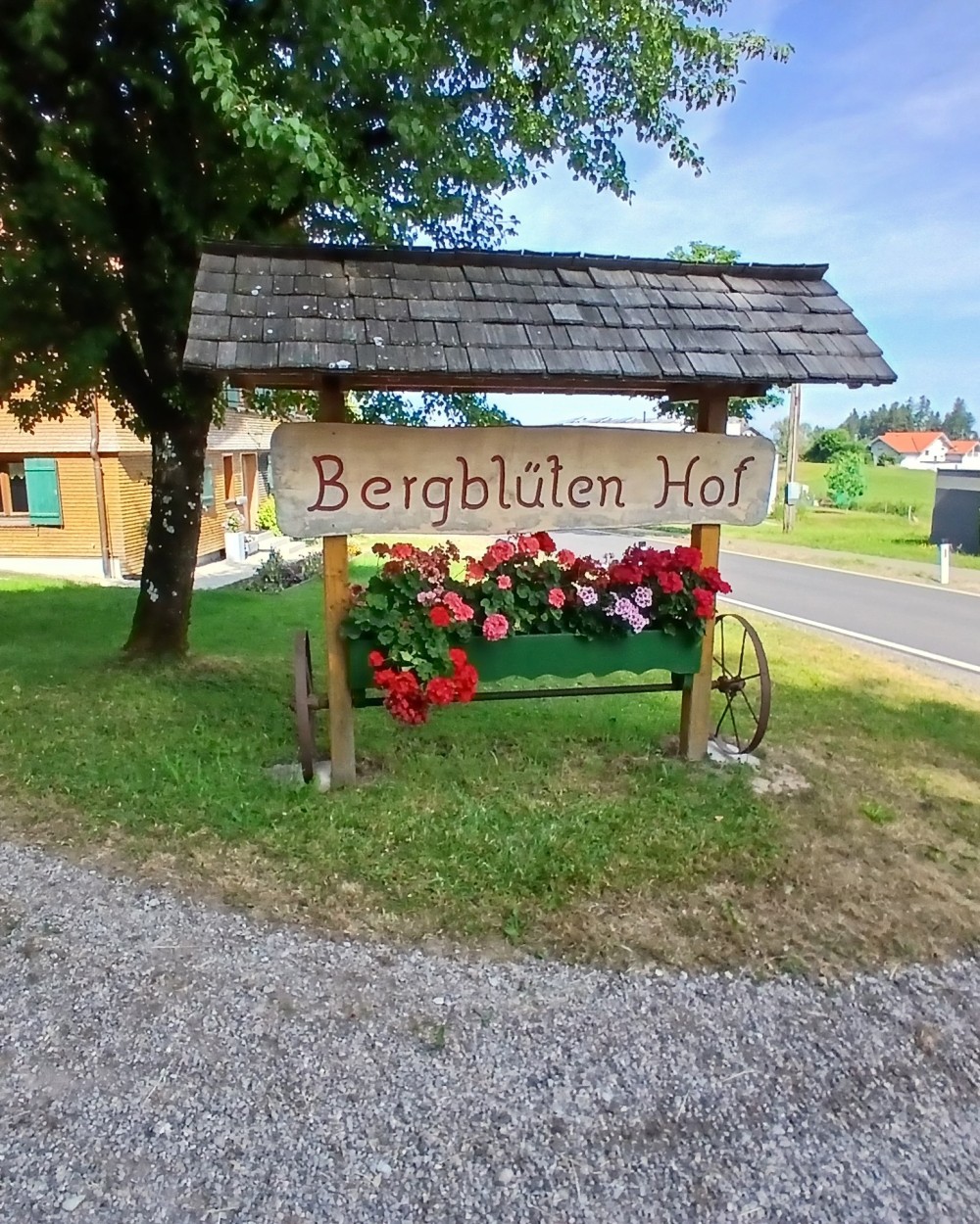 Großes Holzschild des Bergblütenhof - überdacht und darunter rote Geranien gepflanzt. (c) KibeV
