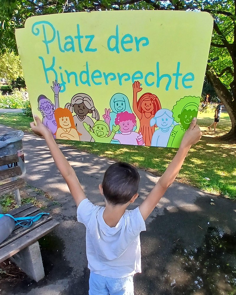 Kind hält ein Schild mit "Platz der Kinderrechte" hoch. (c) KibeV