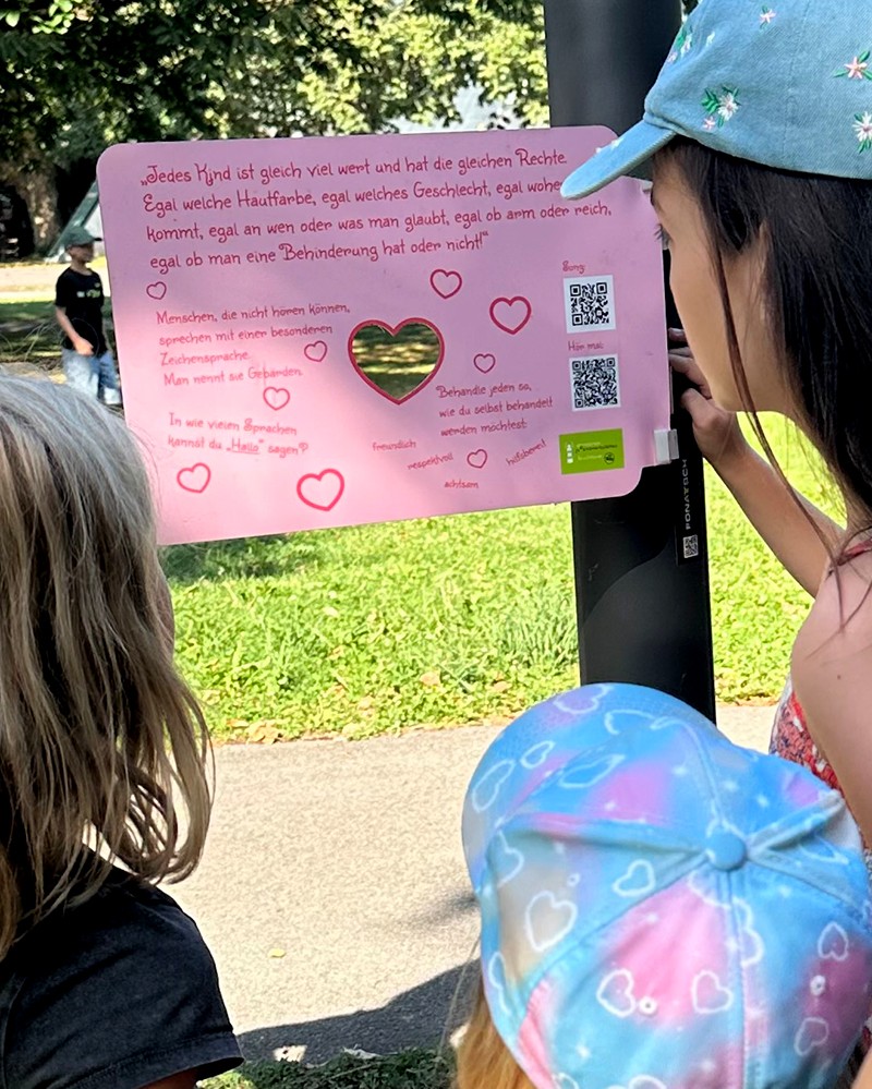 Kinder stehen vor einer Kinderrechte-Tafel und lesen sie aufmerksam durch. (c) KibeV