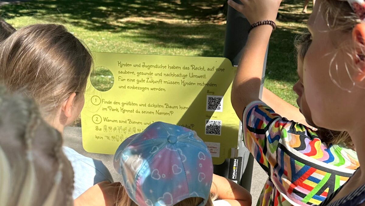 Kinder stehen vor einer Kinderrechte-Tafel und lesen sie aufmerksam durch. (c) KibeV
