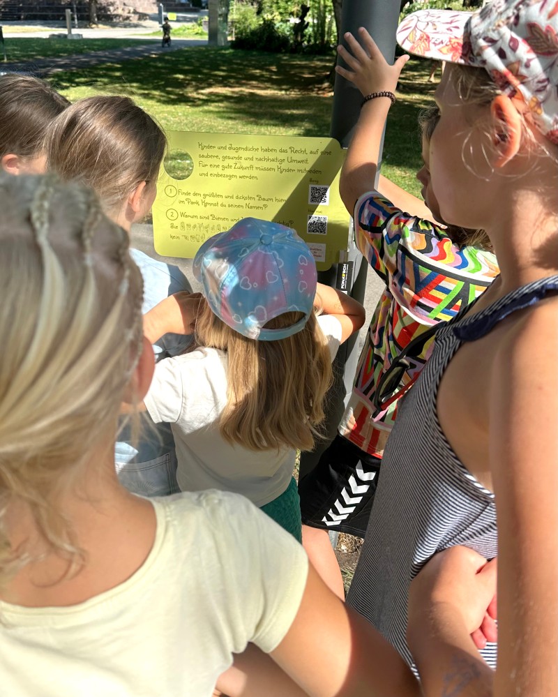 Kinder stehen vor einer Kinderrechte-Tafel und lesen sie aufmerksam durch. (c) KibeV