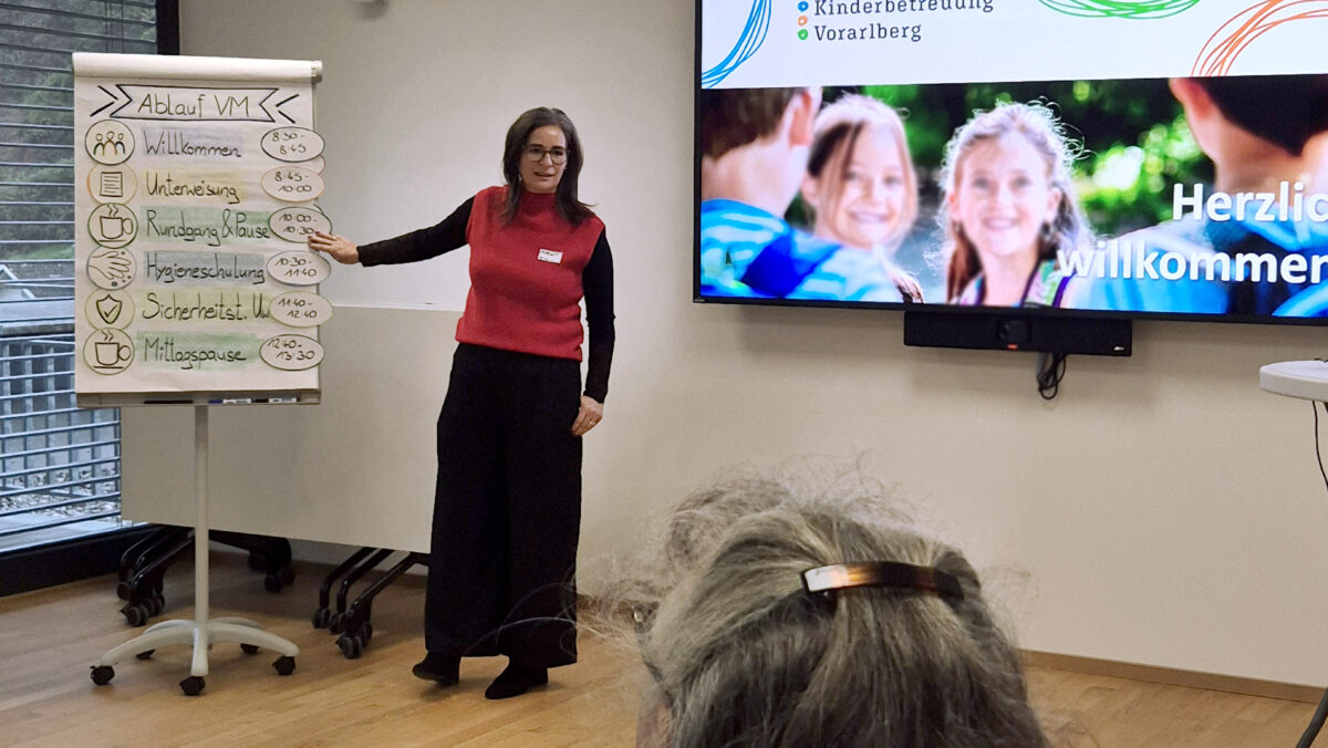 Blick über die Schulter auf Fachbereichsleitung Margot Kernbichler vor einem Flipchart mit den Programmpunkten des Einschulungstages stehend. Rechts von ihr die Starfolie der Präsentation. (c) KibeV