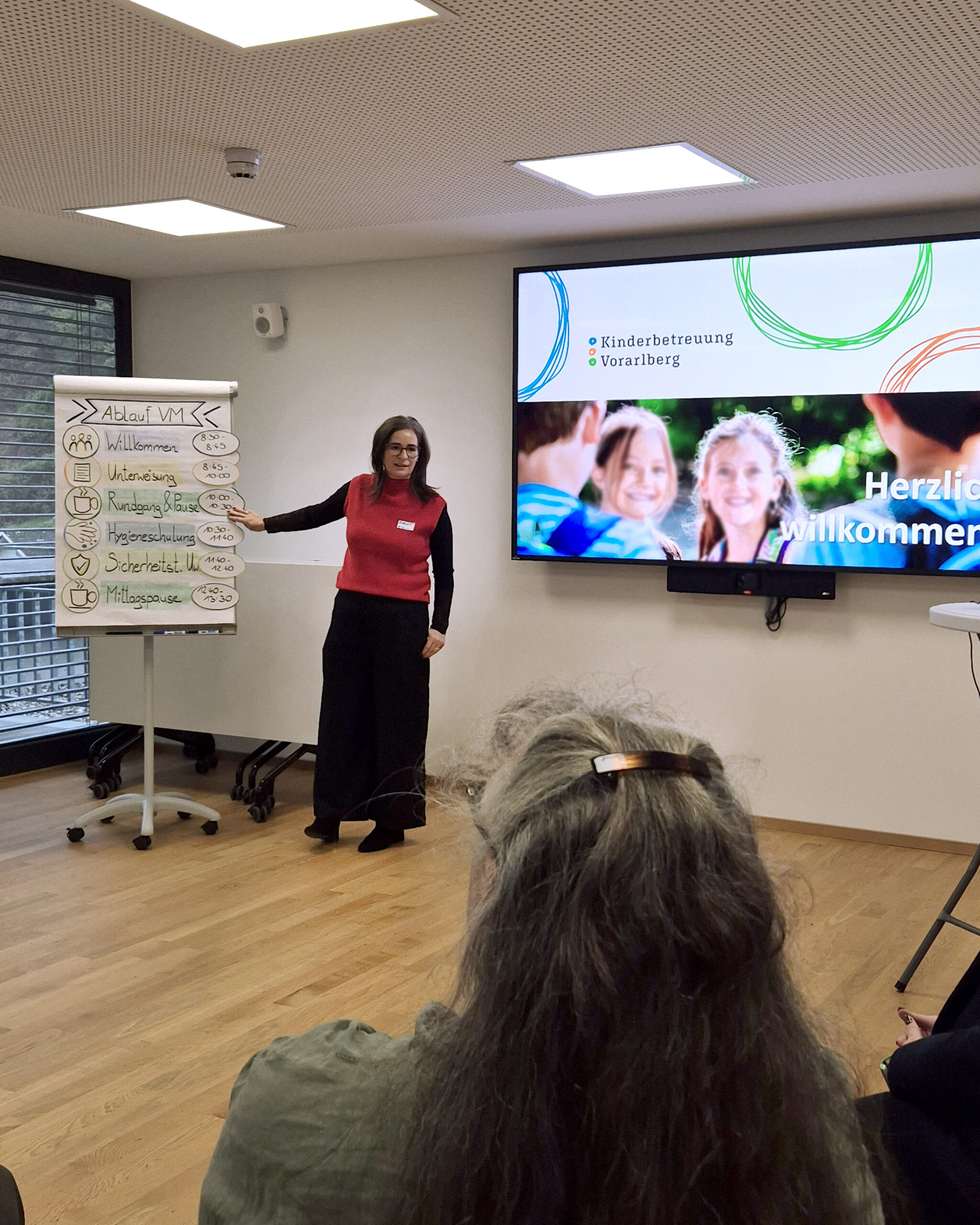 Blick über die Schulter auf Fachbereichsleitung Margot Kernbichler vor einem Flipchart mit den Programmpunkten des Einschulungstages stehend. Rechts von ihr die Starfolie der Präsentation. (c) KibeV