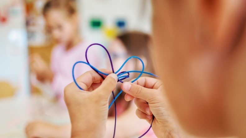 Rückansicht auf Kinderhände, die 2 verschiedenfarbige Schnüre in der Hand halten und damit Schlaufen bilden. (c) KibeV