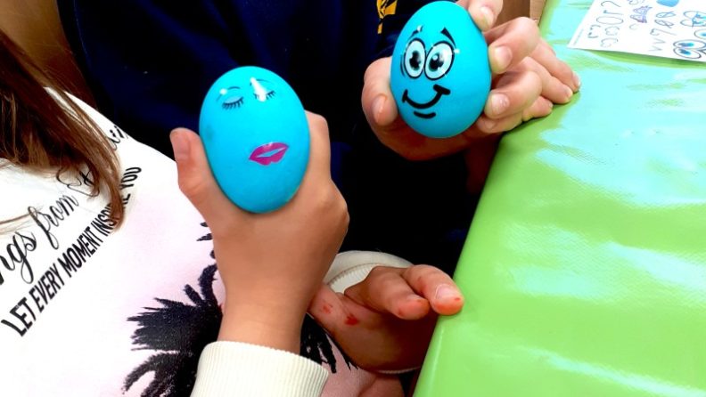 Blick auf die Hände von 2 Kindern, die ihre blauen Ostereier mit lustigem Gesicht präsentieren. (c) KibeV