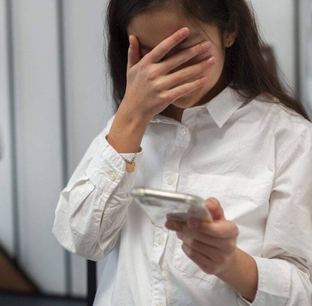 Mädchen weint mit Hand vor dem Gesicht und Handy in der Hand. Im Hintergrund sind verschwommen zwei Kinder zu sehen. (c) Magnific